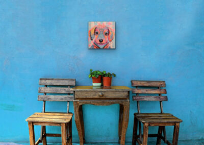 Quadratisches, farbenfrohes Pop-Art-Gemälde eines niedlichen Dackels an einer leuchtend blauen, rustikalen Wand über zwei Holzstühlen und einem kleinen Tisch.