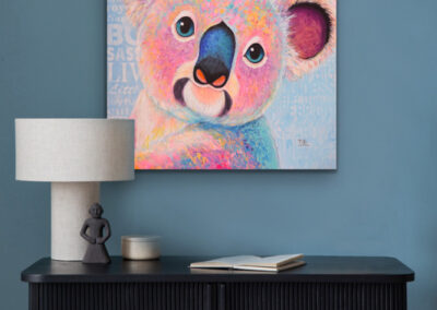 Quadratisches Kunstwerk des bunten Koalas im Pop-Art-Stil an einer dunkelblauen Wand über einem eleganten, schwarzen Konsolentisch mit weißer Tischlampe.