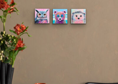 Drei kleine Pop-Art-Tierportraits nebeneinander an einer warmen, braunen Wand über einem Tisch mit einem offenen Buch, einer roten Kaffeetasse und herbstlichen Blumen.