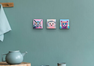 Drei kleine Pop-Art-Tierportraits nebeneinander an einer salbeigrünen Wand über einem Holztisch mit einer blauen Teekanne und Bechern
