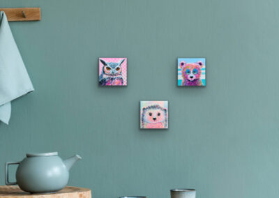 Drei kleine Pop-Art-Tierportraits, als Dreieck angeordnet an einer salbeigrünen Wand über einem Holztisch mit einer blauen Teekanne und zwei Bechern.
