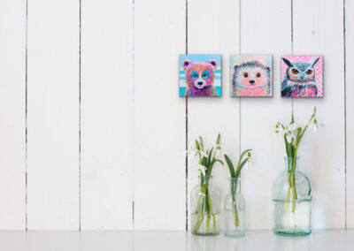 Drei kleine, quadratische, bunte Tierportraits (ein Bär, ein Igel und ein Eule) nebeneinander an einer weißen Holzplanken-Wand über Glasvasen mit grünen Frühlingsblumen.