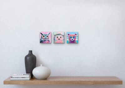 Drei kleine, quadratische Pop-Art-Tierportraits (eine Eule, ein Igel und ein Bär) nebeneinander an einer hellgrauen Wand über einer Holzablage mit Vasen.