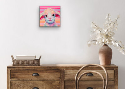 Quadratisches Lamm-Gemälde im Pop-Art-Stil an einer hellen Wand über einer rustikalen Holzkommode mit einer braunen Keramikvase und Trockenblumen.