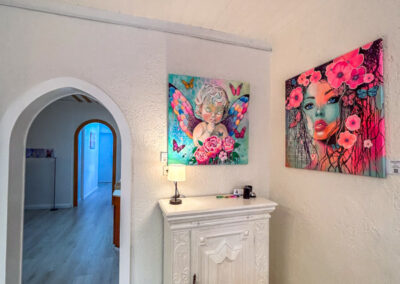 Obere Reihe (v.l.n.r.) 1. Bild ganz links (Flur/Kommode) ALT-Text: Moderne Wandgestaltung im Flur mit farbenfroher Neo-Pop-Art von Silke Timpe. Ein leuchtendes Frauenporträt mit Blumen und ein Schmetterlingskunstwerk harmonieren über einer weißen Vintage-Kommode.