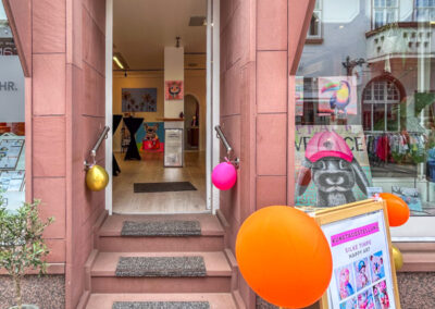 Ankündigung der Kunstausstellung „Happy Art“ von Silke Timpe auf einem hölzernen Aufsteller vor dem Schaufenster – moderne Tierkunst, die Passanten inspiriert.