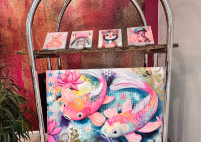 Kreative Kunstpräsentation: Farbgewaltiges Koi-Karpfen-Gemälde von Silke Timpe im Neo-Pop-Art-Stil mit pinken Fischen, dekorativ platziert auf einem silbernen Hotelgepäckwagen vor einer strukturierten Wand.