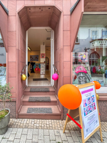 Ankündigung der Kunstausstellung „Happy Art“ von Silke Timpe auf einem hölzernen Aufsteller vor dem Schaufenster – moderne Tierkunst, die Passanten inspiriert.